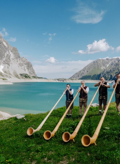 Vier Männer die Alphorn spielen am malerischen Ufer des Lünersee | © Golm Silvretta Lünersee Tourismus GmbH Bregenz, Philipp Schilcher