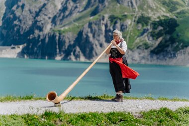 Eine Dame die Alphorn spielt am Ufer des Lünersee | © Golm Silvretta Lünersee Tourismus GmbH Bregenz, Philipp Schilcher