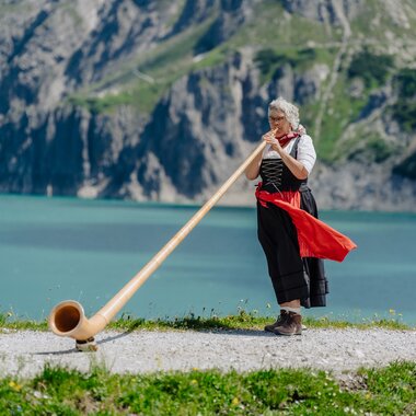 Eine Dame die Alphorn spielt am Ufer des Lünersee | © Golm Silvretta Lünersee Tourismus GmbH Bregenz, Philipp Schilcher