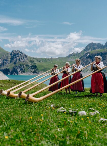 Fünf Damen die Alphorn spielen am malerischen Ufer des Lünersee | © Golm Silvretta Lünersee Tourismus GmbH Bregenz, Philipp Schilcher